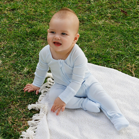 Baby wearing a light blue smart bamboo zipper romper, sitting on a white blanket outdoors, smiling and enjoying playtime on the grass.
