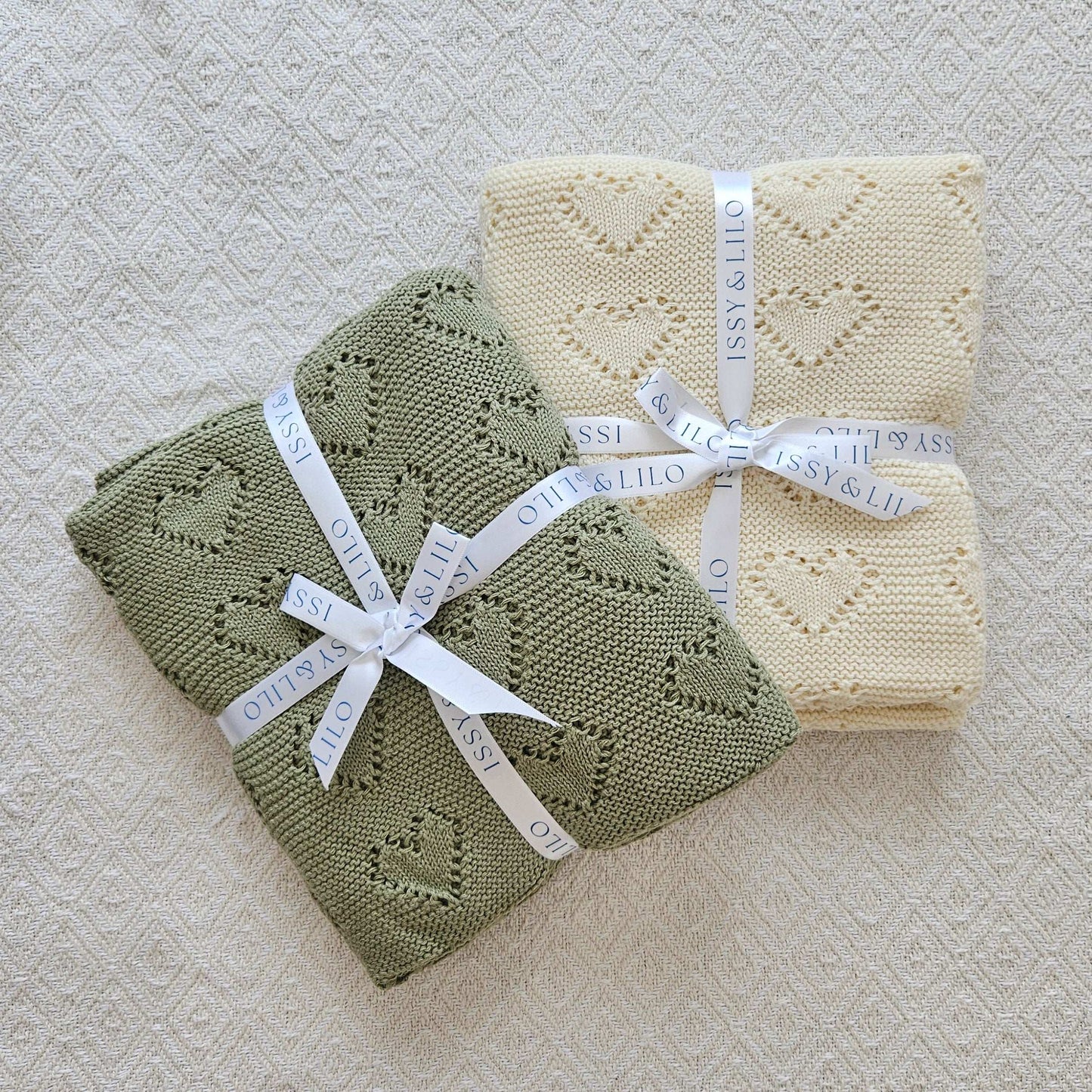 Issy & Lilo organic cotton blankets in sage green and cream, folded and tied with branded ribbon, featuring heart-knit patterns