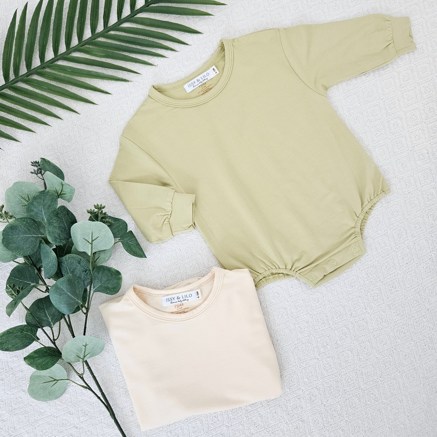 Set of two long sleeve bamboo bodysuits by Issy & Lilo in soft pastel shades of sage green and cream, displayed with greenery accents. Perfect for your baby’s comfort with soft, breathable fabric and easy snap closures.