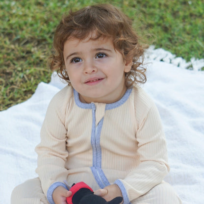 Toddler sitting outdoors on a blanket, wearing a smart organic cotton zipper romper in cream with blue trim.