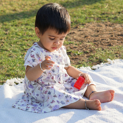 A toddler sits outdoors on a white picnic blanket, dressed in a floral-patterned dress, playing with a red toy. The setting features green grass and natural sunlight, creating a serene and joyful moment.