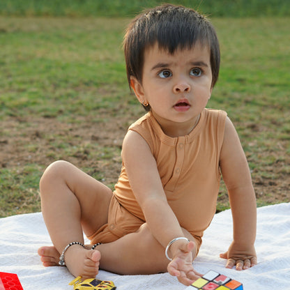 A toddler sits on a white picnic blanket outdoors, dressed in a beige outfit, with a curious expression. The background features green grass, creating a calm and natural atmosphere.