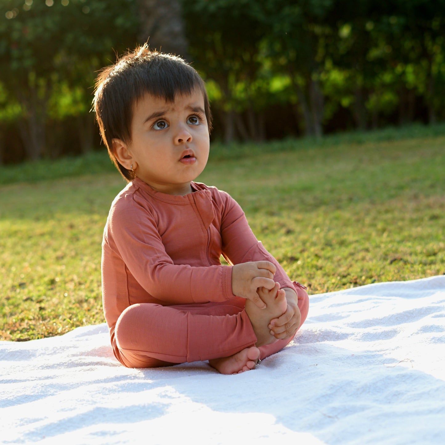 A toddler sits cross-legged on a white blanket outdoors, wearing a rust-colored outfit and gazing upwards with an inquisitive expression. The background features lush green grass and trees, adding a serene and natural vibe.