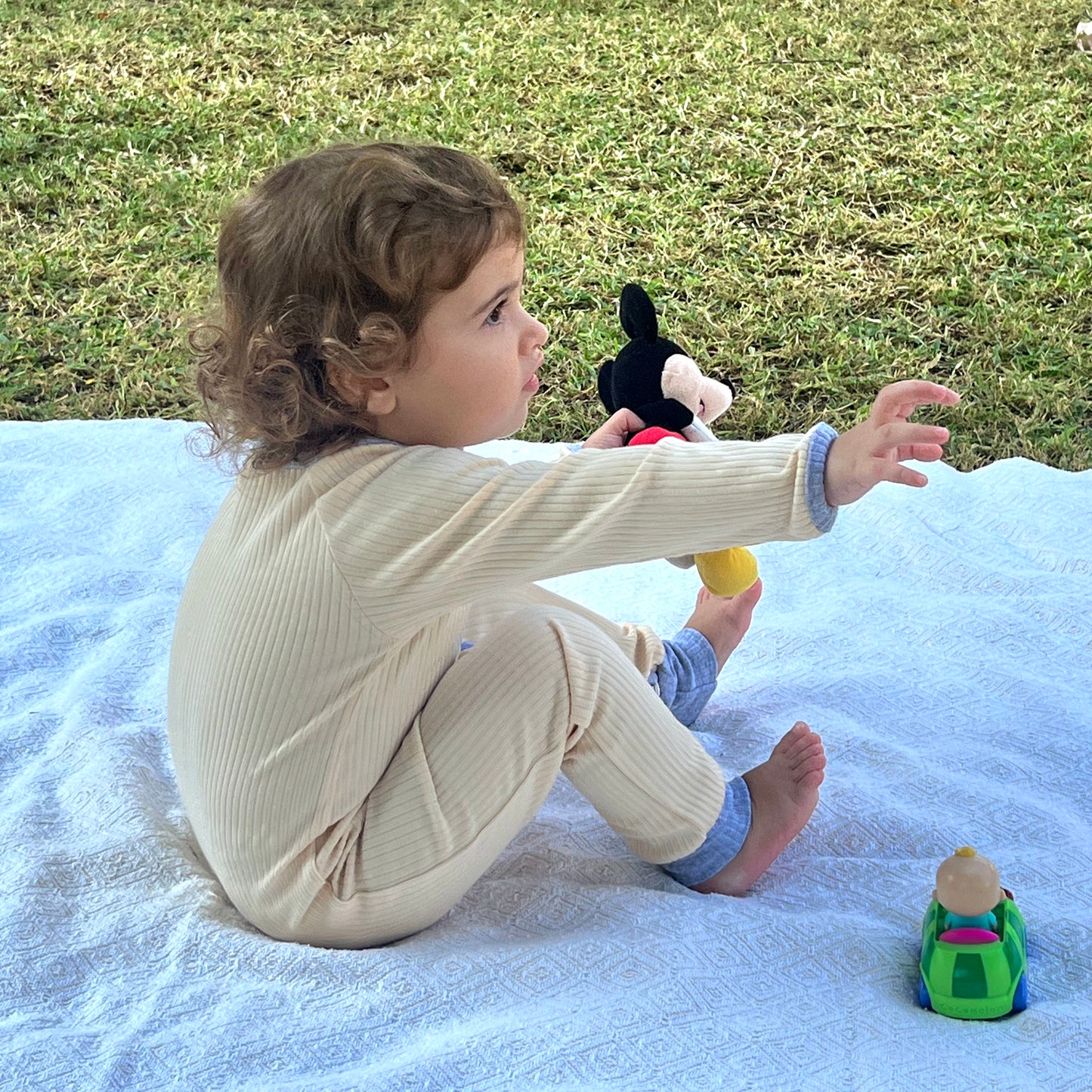 Toddler sitting sideways on a blanket outdoors, dressed in a cream-colored organic cotton zipper romper with blue accents, playing with a small toy.