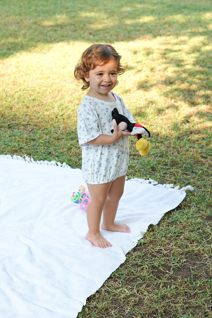 Toddler standing on a white blanket outdoors, holding a toy, wearing an Issy & Lilo light blue organic cotton baby bodysuit