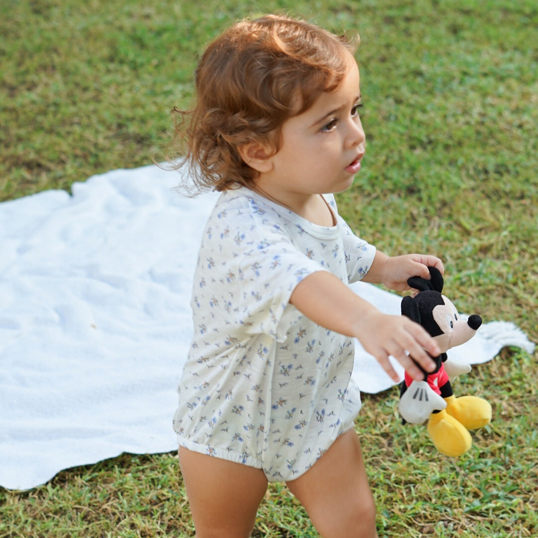 Toddler walking on grass with a toy in hand, wearing an Issy & Lilo light blue organic cotton baby bodysuit, with a white blanket in the background