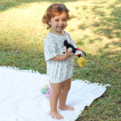 Toddler standing on a white blanket outdoors, holding a toy, wearing an Issy & Lilo light blue organic cotton baby bodysuit