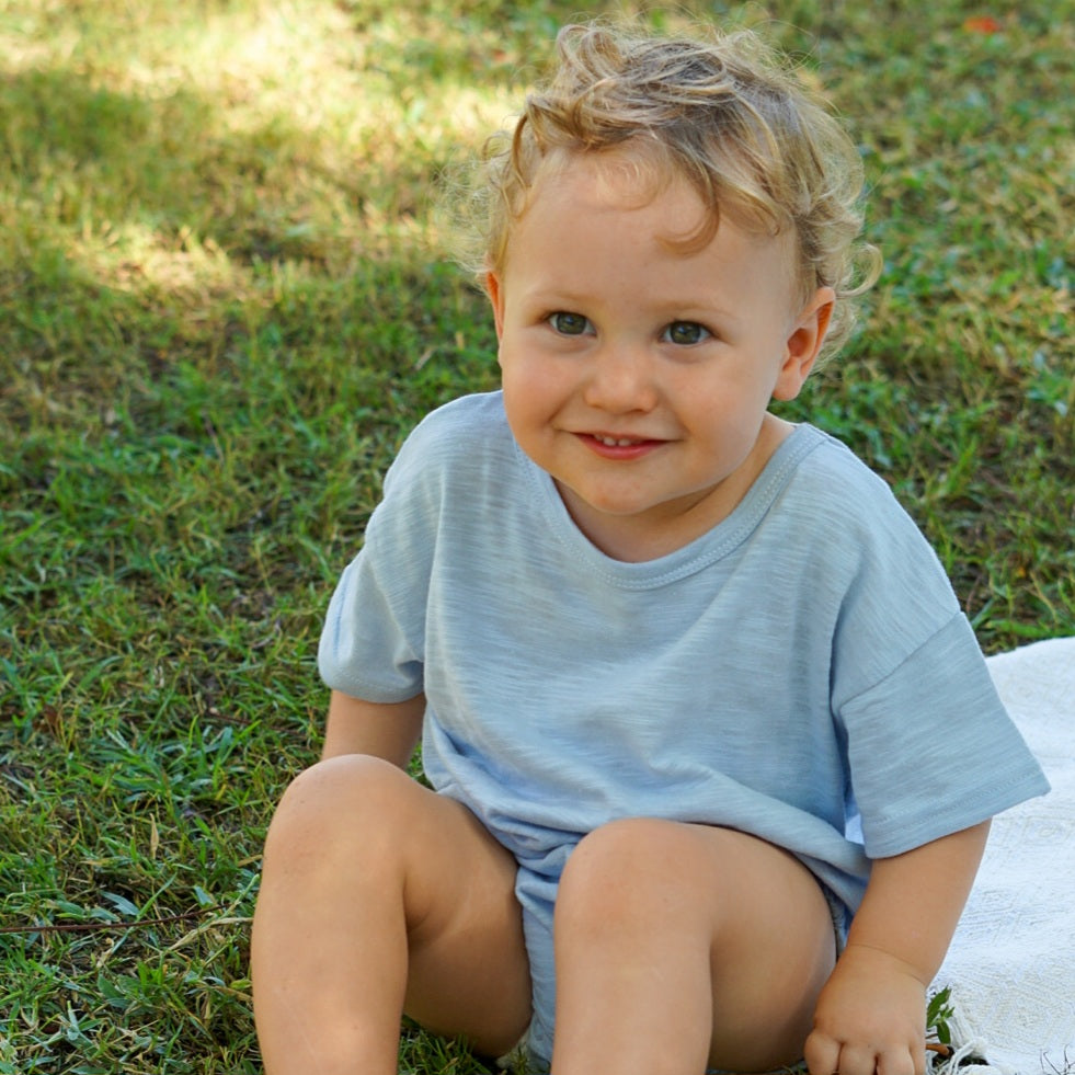 Toddler smiling outdoors, wearing an Issy & Lilo light blue organic cotton t-shirt, sitting on grass